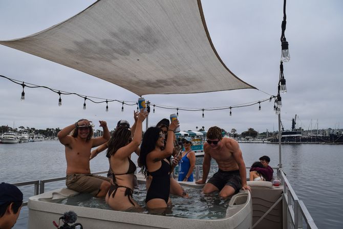 Friends splashing in a hot tub on a pontoon at a marina, raising canned drinks under a fabric sunshade and string lights with boats and a cloudy sky beyond.