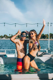 Two adults in swimwear dancing in a boat hot tub with a cocktail, sunglasses and ocean with palm-lined coastline on a sunny summer day
