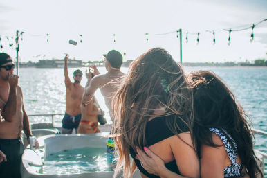 Two women hugging on a sunlit boat deck party, friends in swimwear by a small hot tub, string lights overhead and the ocean coastline sparkling in the background