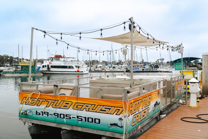 Floating hot-tub boat moored at a marina dock under string lights and a shade sail, with cushioned seating and sailboats and ferries in the harbor behind.