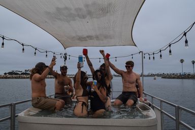 Friends partying in a hot tub on a pontoon boat at a coastal harbor marina, raising cups under string lights with palm trees on an overcast day.