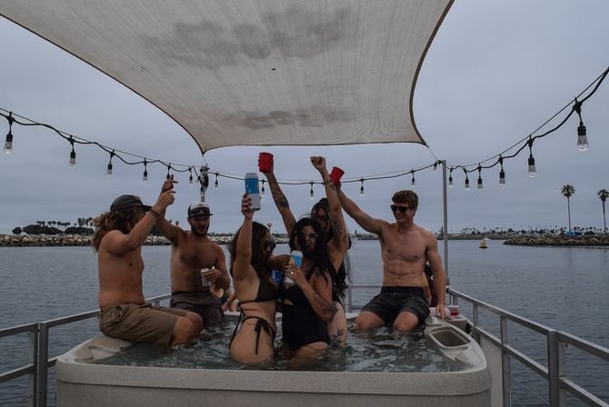 Friends partying in a hot tub on a pontoon boat at a coastal harbor marina, raising cups under string lights with palm trees on an overcast day.