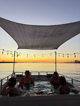 Group relaxing in a boat hot tub at sunset beneath a stretched shade and Edison string lights, calm bay water with palm-tree-lined horizon and warm golden sky