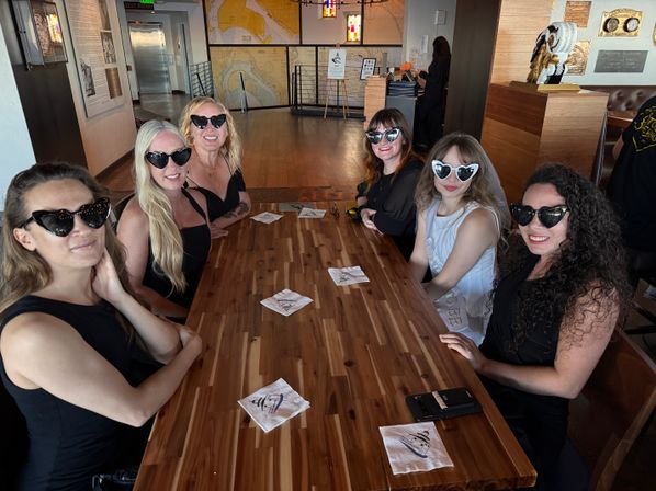 Group of six women wearing heart-shaped sunglasses around a long wooden table in a harbor-themed restaurant — smiling bachelorette party with bride-to-be in a white sash and nautical maps on the wall.