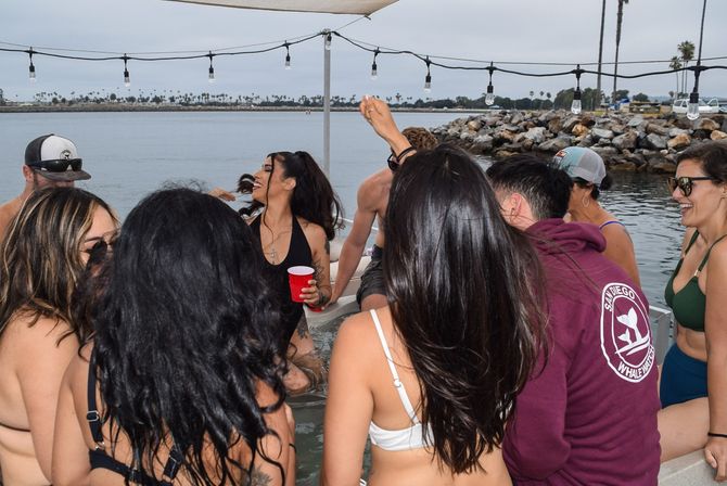 Group of friends in swimsuits laughing and chatting at a lively boat party by a rocky harbor with palm trees in the distance, string lights overhead and a red party cup in hand.