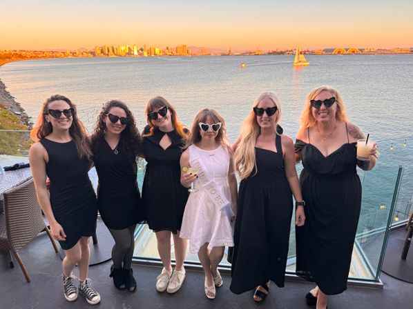 Six women in black dresses and a bride-to-be in a white sash wearing heart-shaped sunglasses, posing on a waterfront rooftop with a sailboat and the San Diego skyline glowing at sunset over the bay