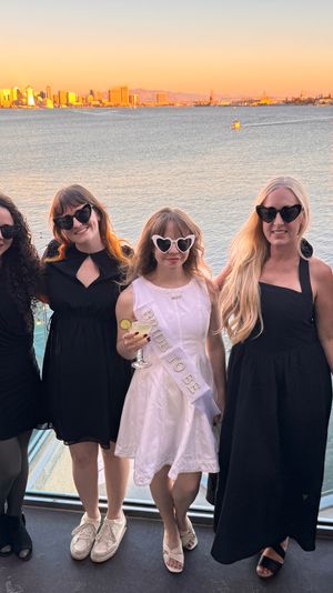 Six women in black dresses and a bride-to-be in a white sash wearing heart-shaped sunglasses, posing on a waterfront rooftop with a sailboat and the San Diego skyline glowing at sunset over the bay