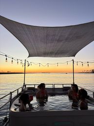 People relaxing in a hot tub on a boat at sunset over a calm bay, string lights and shade sail silhouetted against a colorful sky with distant palm trees