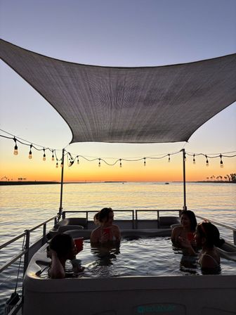 People relaxing in a hot tub on a boat at sunset over a calm bay, string lights and shade sail silhouetted against a colorful sky with distant palm trees