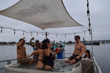 Friends laughing and holding drinks in a hot tub on a pontoon boat at a marina, under a shade sail and string lights.