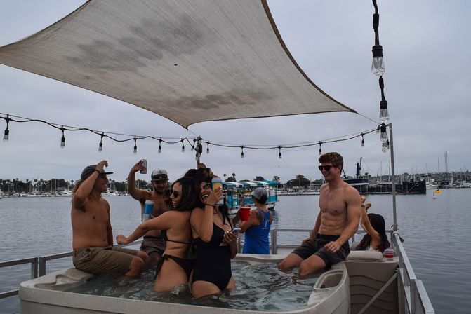 Friends laughing and holding drinks in a hot tub on a pontoon boat at a marina, under a shade sail and string lights.