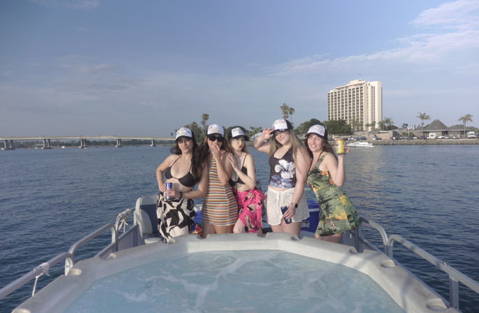 Five friends in swimsuits and matching trucker hats posing with drinks on a pontoon boat’s hot tub, waterfront skyline and bridge in the background