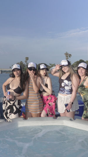 Five friends in swimsuits and matching trucker hats posing with drinks on a pontoon boat’s hot tub, waterfront skyline and bridge in the background