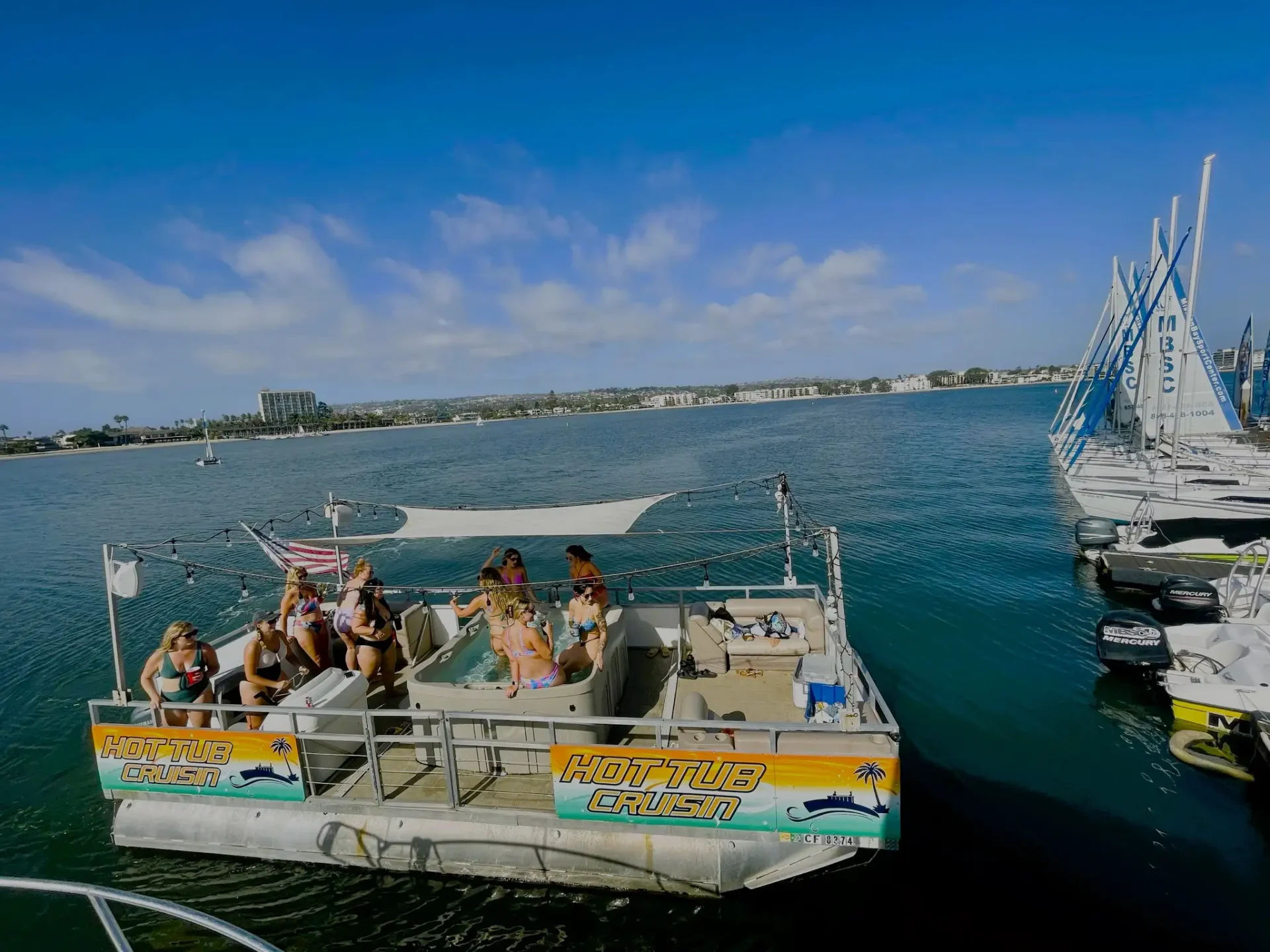 Sunny hot-tub pontoon boat with people enjoying a boat party in a calm coastal bay marina with sailboats docked nearby.