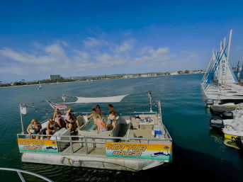 Sunny hot-tub pontoon boat with people enjoying a boat party in a calm coastal bay marina with sailboats docked nearby.