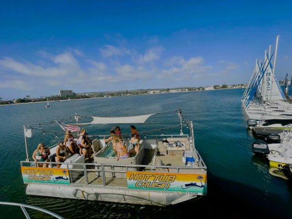 Sunny hot-tub pontoon boat with people enjoying a boat party in a calm coastal bay marina with sailboats docked nearby.