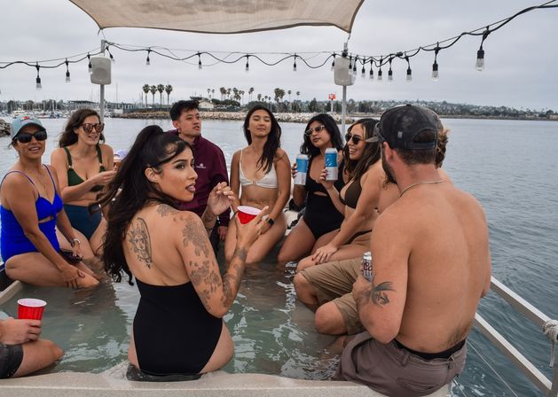 Group of friends in swimsuits sipping drinks and chatting in a pontoon boat's shallow wading area under string lights, with a palm-lined marina and cloudy coastal horizon in the background.