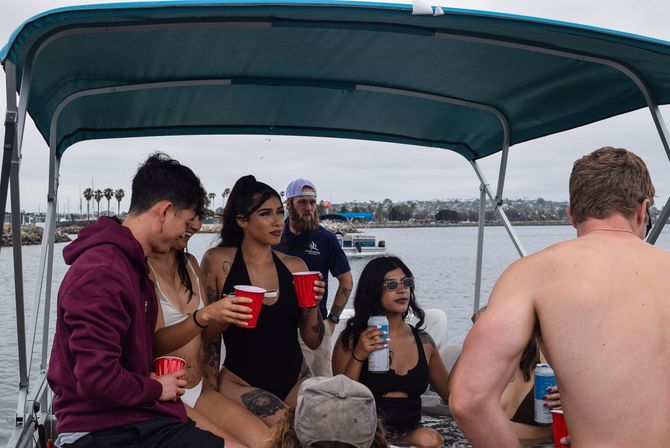 Group of friends on a pontoon boat under a teal canopy at a coastal marina with palm trees, holding red cups and cans on an overcast day.