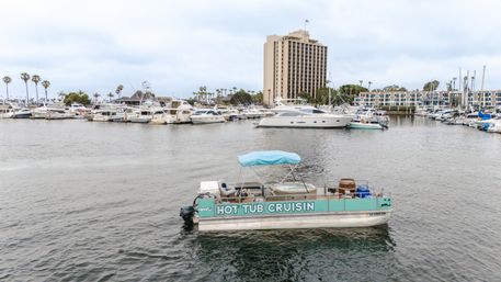 Pontoon labeled 'Hot Tub Cruisin' with a covered hot tub and seating floating in a busy marina, surrounded by yachts, palm trees and a waterfront high-rise under a cloudy sky.
