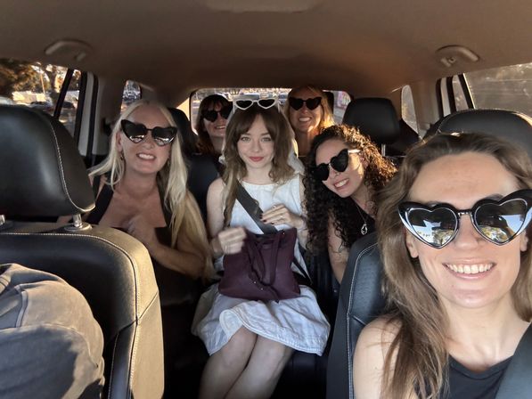 Group selfie of six friends inside a car wearing heart-shaped sunglasses, smiling in warm sunset light — playful road-trip vibe.