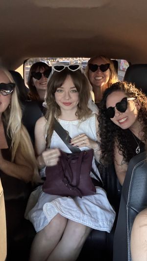 Group selfie of six friends inside a car wearing heart-shaped sunglasses, smiling in warm sunset light — playful road-trip vibe.