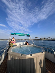 Three people relaxing on a boat with a bubbling hot tub in a calm coastal marina, palm-lined shoreline and sailboats under a bright blue sky streaked with wispy clouds.