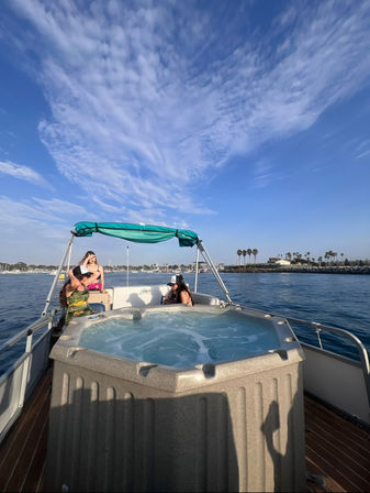 Three people relaxing on a boat with a bubbling hot tub in a calm coastal marina, palm-lined shoreline and sailboats under a bright blue sky streaked with wispy clouds.