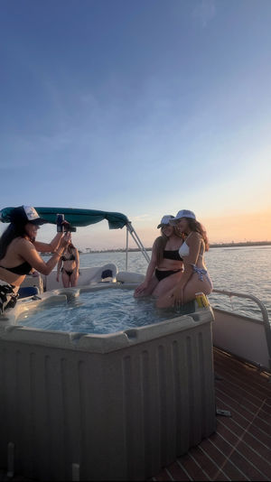 Friends in swimsuits on a pontoon boat hot tub posing for photos at sunset over a calm coastal bay