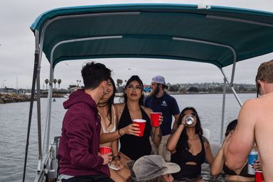 Young adults on a party boat in a coastal bay with palm trees and a marina in the background, holding red cups and drinks under a teal canopy.