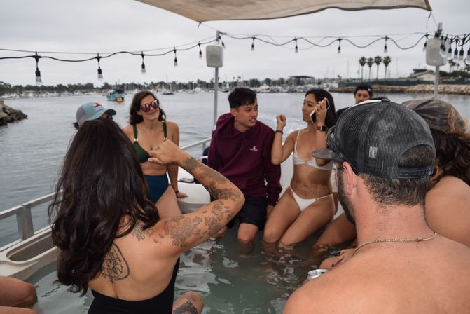 Group of people socializing in a boat hot tub near a coastal marina — swimwear and a hoodie, visible tattoos, string lights above, boats and palm trees in the harbor under cloudy skies.