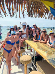 Group of women in swimsuits at a sunny tropical boat bar under a thatched roof, celebrating a bachelorette with the bride in white wearing a 'BRIDE' hat, shallow turquoise ocean and anchored boats in the background