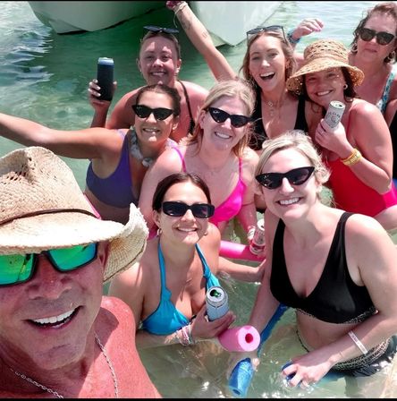 Smiling group of friends in swimsuits and sunglasses standing in shallow turquoise water by a boat, holding canned drinks and pool noodles for a sunny selfie.