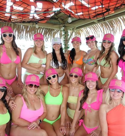 Group of women in neon bikinis and matching pink caps smiling and posing under a thatched beach cabana