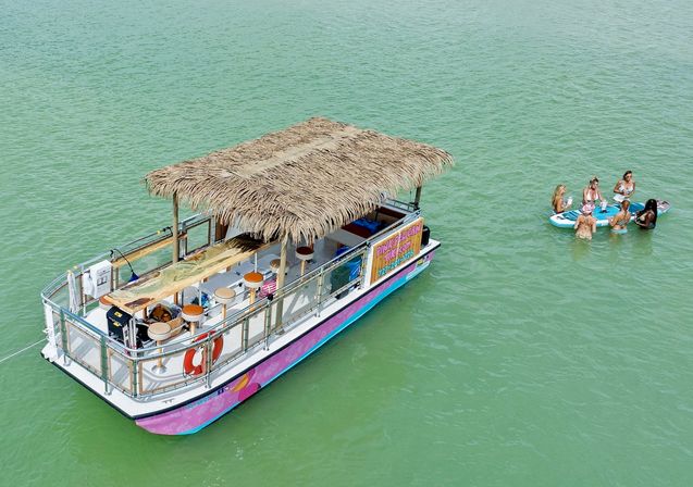 Tiki-style pink pontoon boat with a thatched roof and bar seating anchored in green coastal water, with a small group gathered on a paddleboard nearby for a relaxed boat-and-paddleboard party scene.