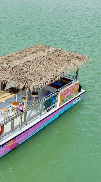 Tiki-style pink pontoon boat with a thatched roof and bar seating anchored in green coastal water, with a small group gathered on a paddleboard nearby for a relaxed boat-and-paddleboard party scene.