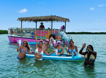 Six women in bikinis holding drinks on a paddleboard beside a colorful pink tiki-style party boat with a thatched roof in shallow tropical bay