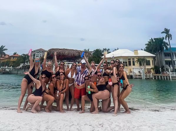 Cheerful group of friends in swimsuits posing on a sandy beach by a tropical waterfront with palm trees, a thatched tiki hut and waterfront homes, holding colorful drinks and celebrating.