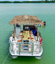 Pontoon party boat with a thatched tiki roof and wooden bar table, anchored in shallow turquoise lagoon near mangrove islands with swimmers floating nearby.