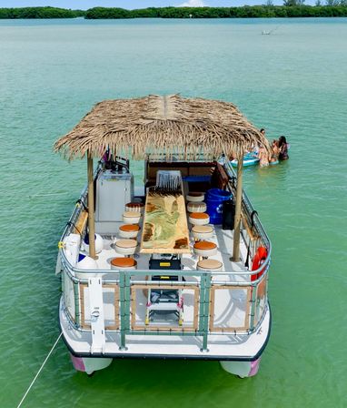 Pontoon party boat with a thatched tiki roof and wooden bar table, anchored in shallow turquoise lagoon near mangrove islands with swimmers floating nearby.