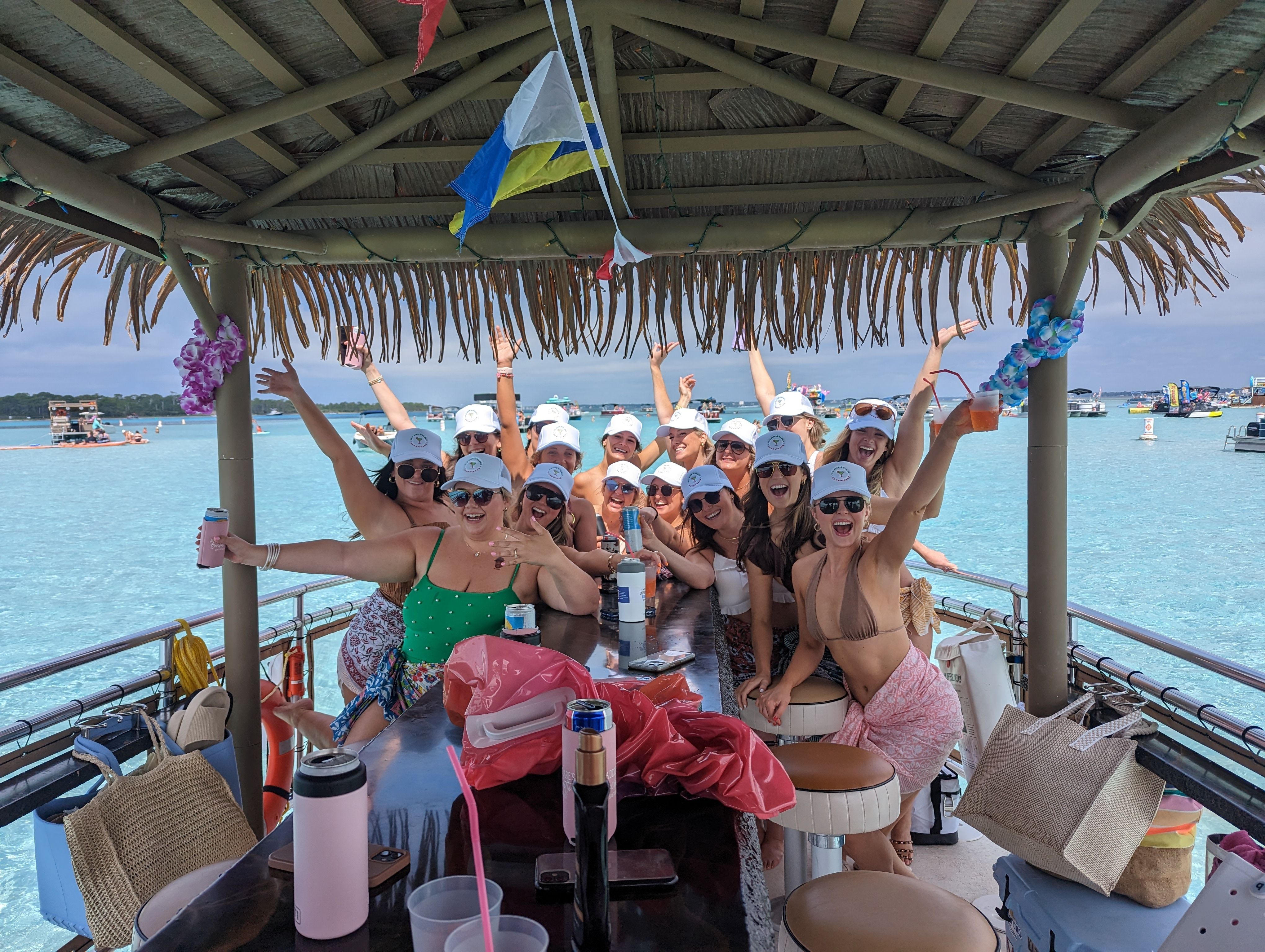 Boat party with a cheering group of women in swimsuits and white caps under a thatched tiki-roof bar, holding drinks over clear turquoise water with boats in the background