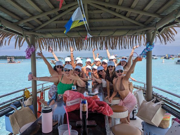 Boat party with a cheering group of women in swimsuits and white caps under a thatched tiki-roof bar, holding drinks over clear turquoise water with boats in the background
