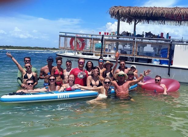 Group of friends enjoying a sunny boat party in shallow turquoise coastal water, posing on a large paddleboard and inflatable tubes beside a thatch-roof party boat.
