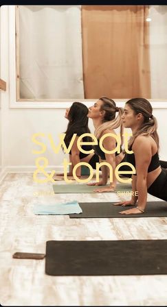 Three women in a bright indoor fitness studio practicing upward-facing dog on yoga mats, with yellow “sweat & tone” text overlay.