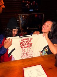 Two smiling bar patrons holding a white t-shirt that reads 'I SURVIVED THE BIGFOOT BEER BOOT CHALLENGE' over a wooden bar counter in a dim, rustic pub.