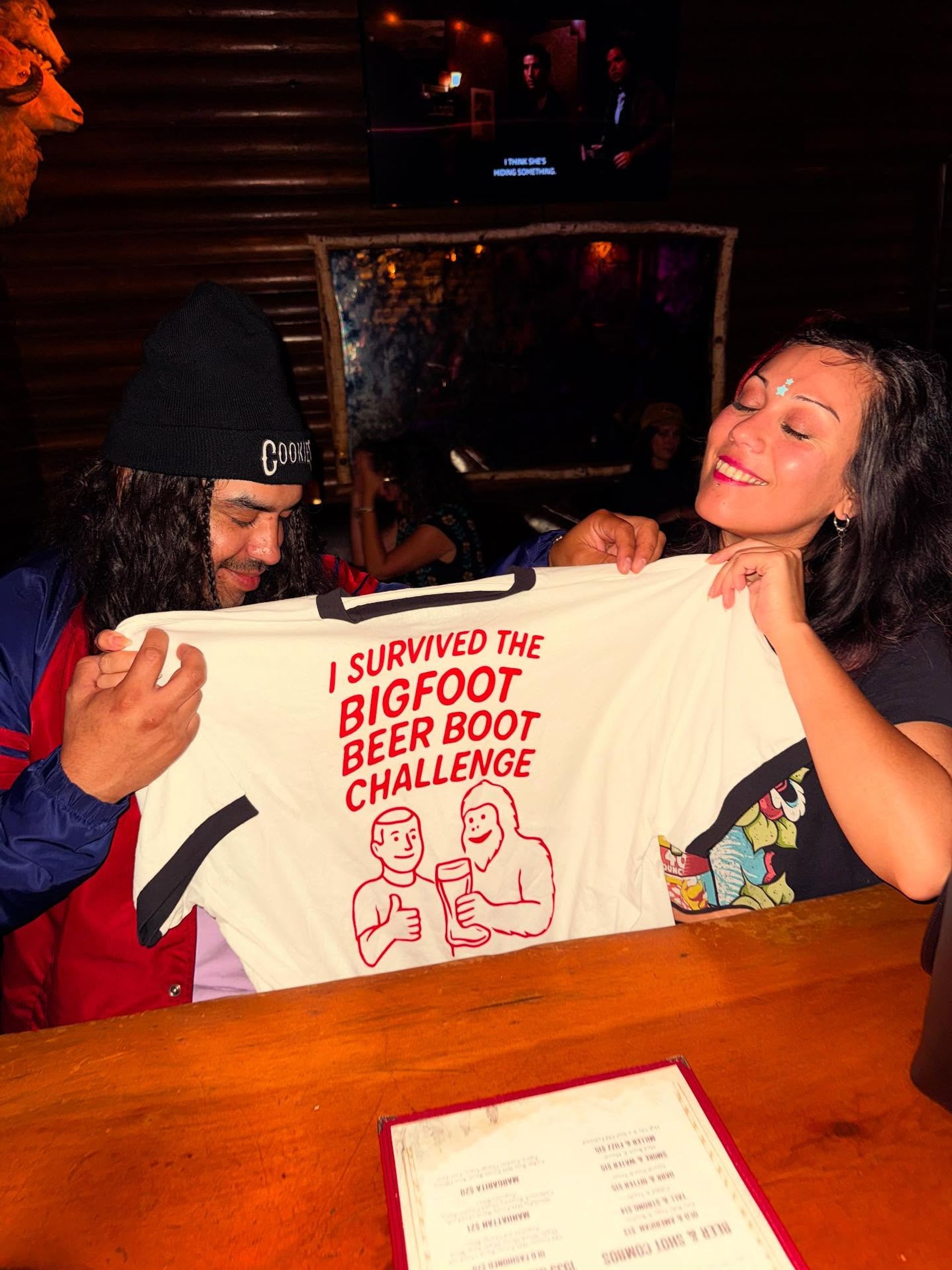 Two friends at a cozy wood-paneled bar holding a white T‑shirt that reads "I survived the Bigfoot Beer Boot Challenge," smiling over a wooden table with a menu — fun pub celebration scene.
