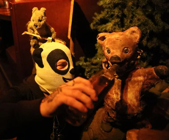 Masked figure in a panda ski mask pretends to feed a bottle to a vintage teddy bear puppet in a dim, amber-lit indoor scene with other stuffed animals.