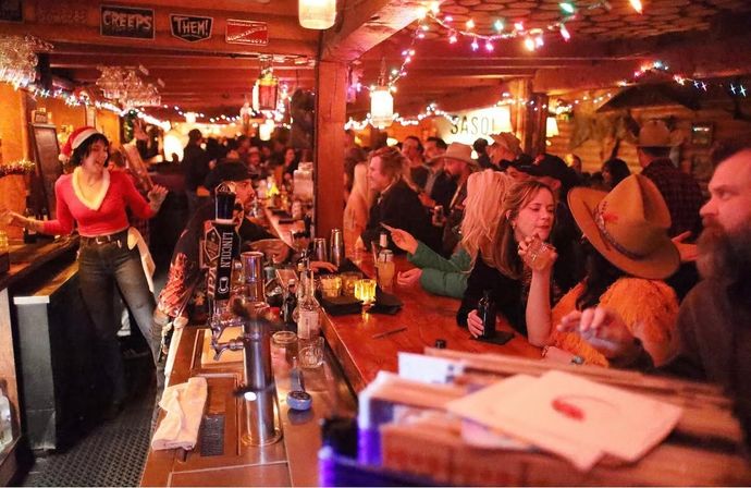 Festive crowded rustic bar with wooden beams and string lights, patrons in cowboy hats and a bartender in a Santa hat serving cocktails at a long wooden bar.