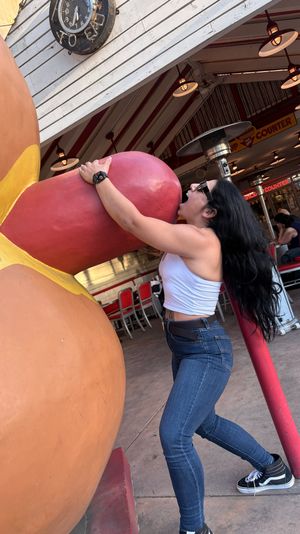 Person in white tank top and jeans playfully pretends to bite a giant red hot dog sculpture outside a retro-style diner with striped awning, red chairs and outdoor seating.