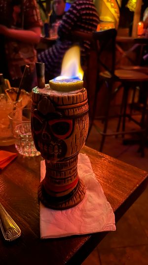 Flaming tiki skull mug cocktail topped with a burning lime on a napkin in a dimly lit tropical-style bar