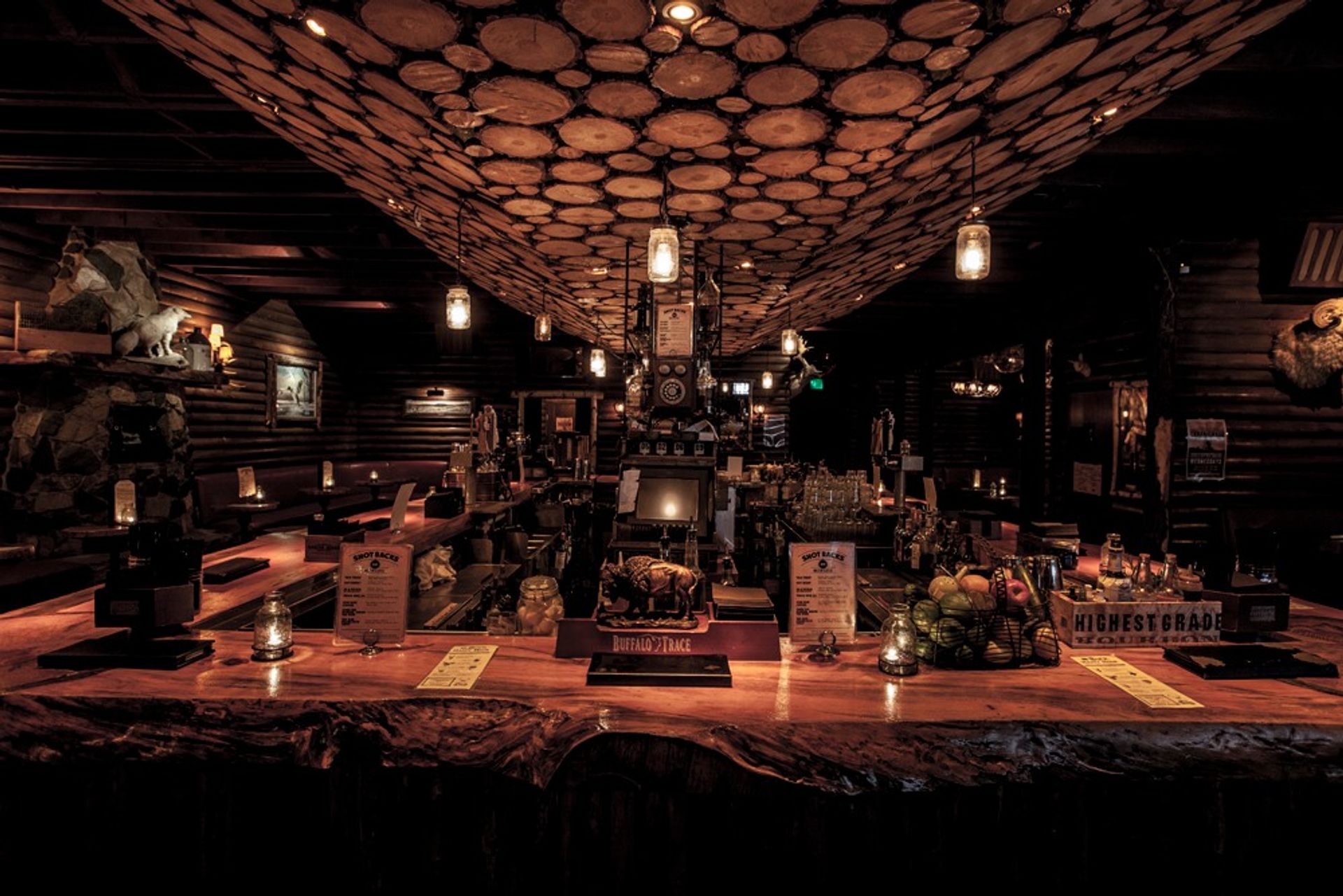 Cozy rustic log-cabin bar interior with a live-edge wooden bar, wood-slice ceiling pattern, hanging mason-jar lights, stone fireplace and dim amber ambient lighting